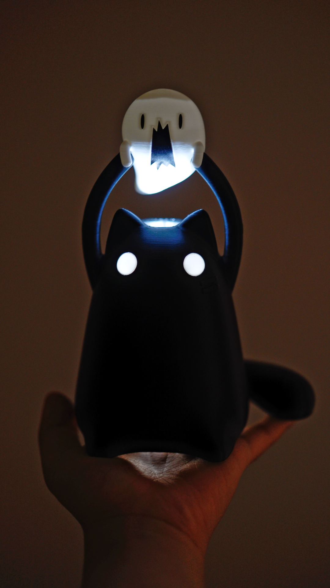 A cat shaped LED light is in the dark, its eyes are glowing white and the mini ghost it is holding above its head is also illuminated