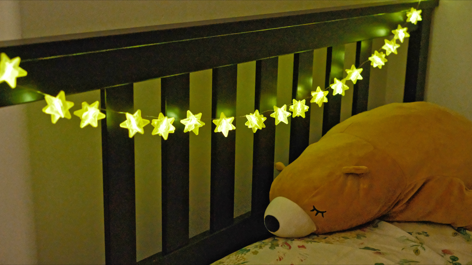 A string light of yellow star shapes are hanging on a bed frame where a stuffed animal bear is placed on top of the pillow on the bed. The string light is shining brightly in the dim room.